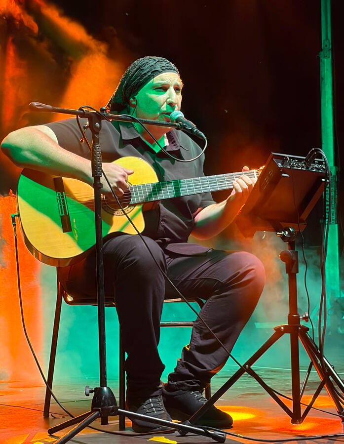 Peppone Ciauda che canta davanti a un microfono su un palco, mentre suona una chitarra acustica. Il musicista indossa un copricapo scuro e una camicia scura. È ripreso di profilo con una mano alzata in un gesto espressivo; l'illuminazione di scena crea un